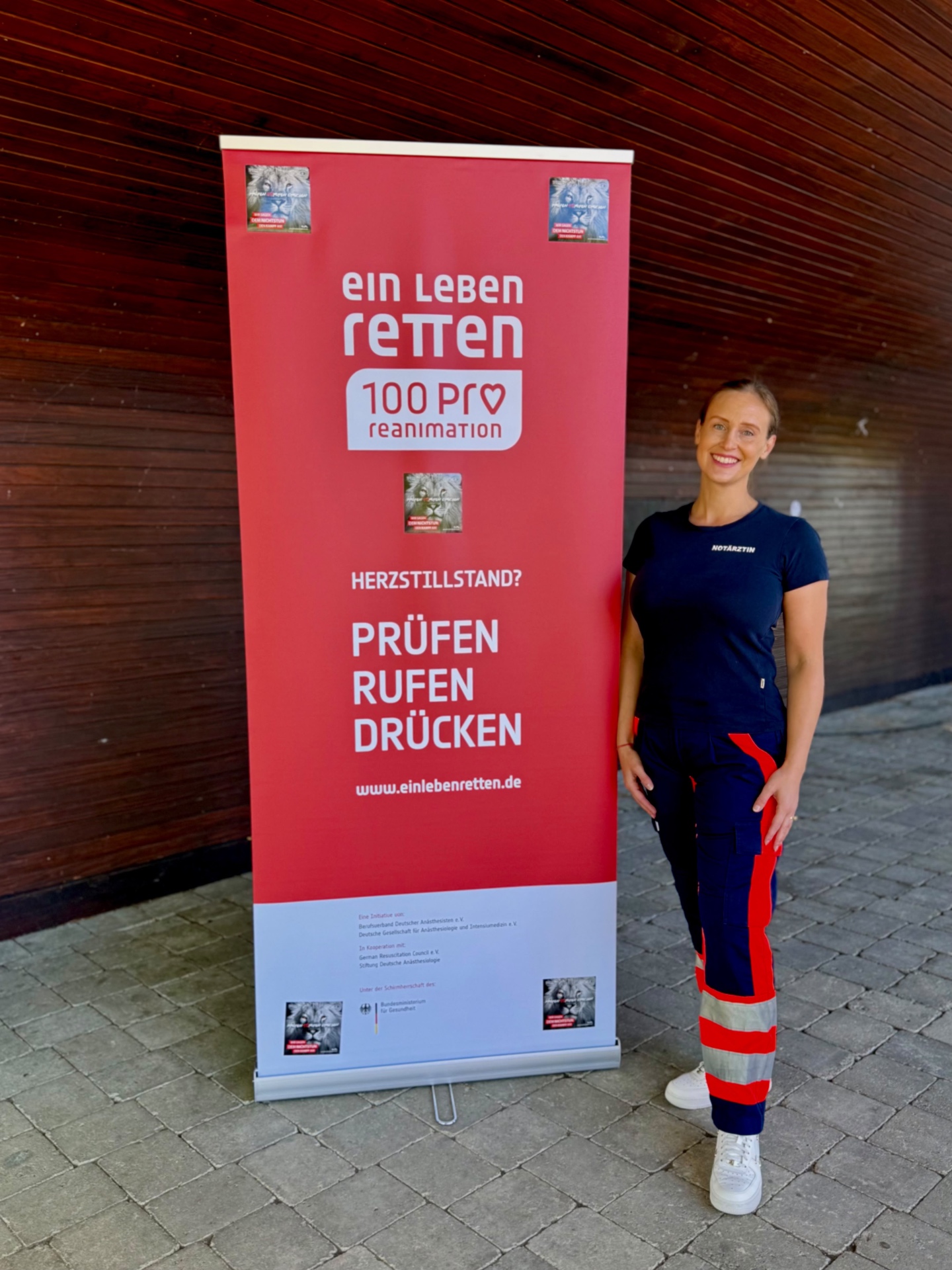 Anna Vormeyer in Notarzt-Einsatzkleidung neben dem Banner „Ein Leben retten – Prüfen, Rufen, Drücken“